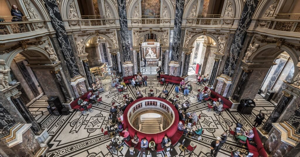 Ornate museum interior with marble columns, sculptures, patterned floors, and visitors seated around a central staircase.